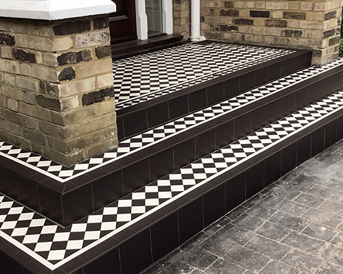Black and white chequerboard tiles on a set of exterior entrance steps, framed with old brick walls with white painted coping stone on sides.