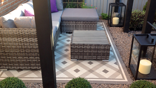A traditional mosaic pattern in a modern grey tone colour palette makes a stunning outdoor patio floor. Wicker sofa, chair and table, and two electric candles, complete the outdoor setting.