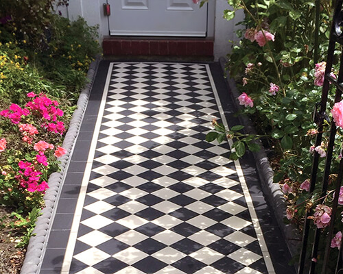 Traditional four inch, black and white ceramic outdoor path tiles, with rose bushes to sides, leading from metal gate to front door.