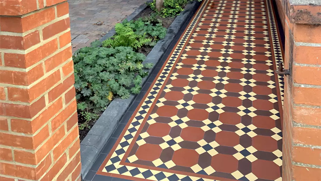 A reproduction Edwardian tiled path using octagon, hexaon and square shapes in red, cream, black and brown colours. Our view of the path is between two brick piers. To the right, curved black bricks finish a bed of shrubs. Beyond this, a stone-brick driveway.