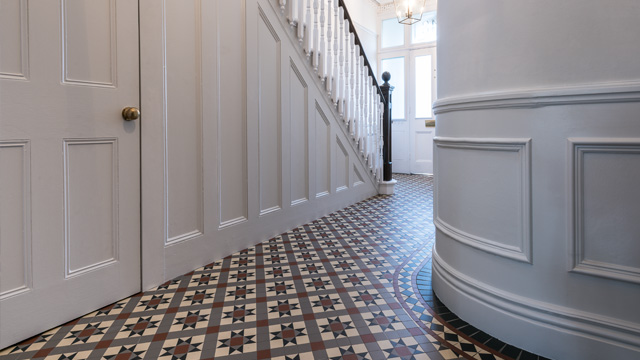 Large hallway with panelling on walls and staircase, featuring traditional curved wall. Period style Victorian floor tiles in red, grey, black and ivory colours.