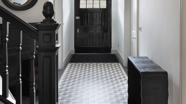 A Victorian property with a wide hallway has modern grey-toned mosaic floor tiles. There is light reflecting off the tiles. To the rear of the picture a large black front door with dark coir mat in front. To the left, a staircase with dark wooden carved banister and newel post. On the right a dark wood side-table.