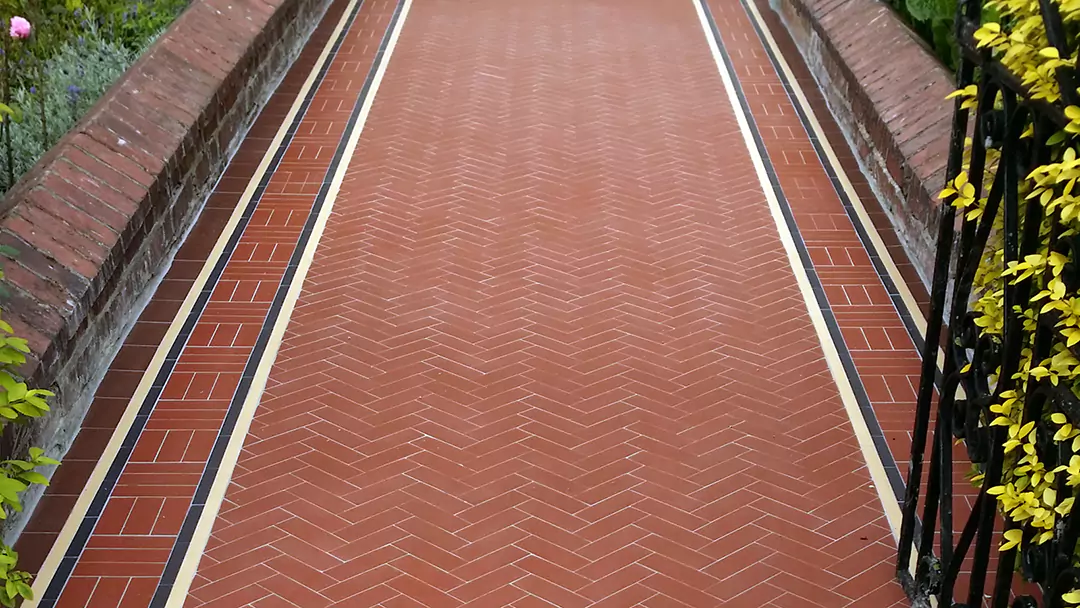 Edwardian red herringbone tiles are fitted to a wide entrance path. To the left and right, small brick walls, finished with a triangular peak, hold back planted shrubs and flowers. To the right, a black wrought-iron gate with the yellow-green leaves of a hedge behind it.