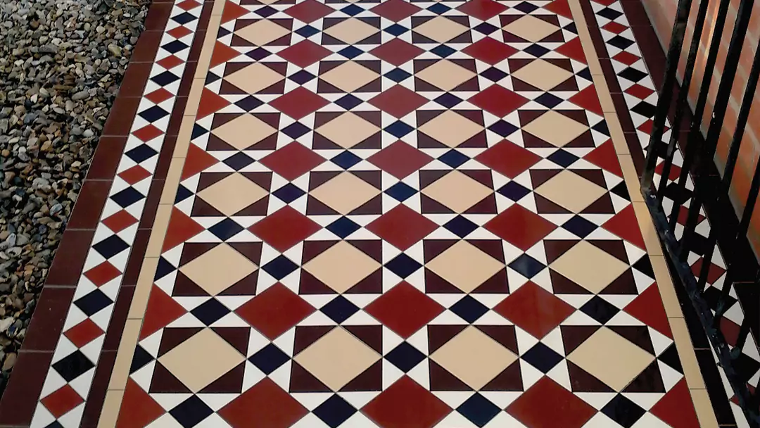 A colourful geometric Edwardian path tile design using black, white, brown, red and cream tiles, framed by a diamond-mmotif border on both sides. To the right, an open metal gate and red-brick wall. To the right, grey pebbles.