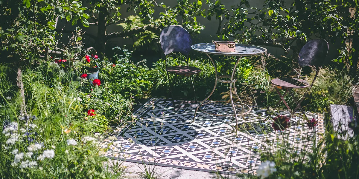A garden patio has decorative Victorian mosaic tiles in a traditional pattern. There are two old metal chairs and a metal table with a shiny copper pot on top. The garden foliage surrounds the patio, it is bright green, lit by strong sunlight. There are many different flowers on display.