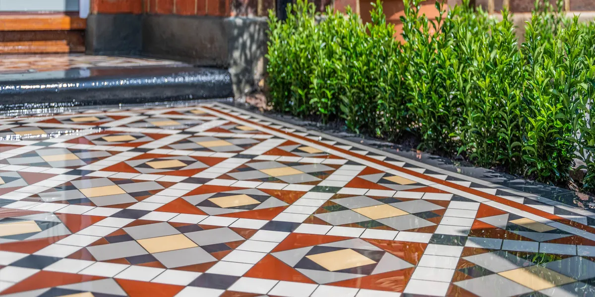 A closer view of a red, grey and yellow coloured Edwardian mosaic pathway, with shrubs planted to the sides. A small stone step painted black leads to the porch. The path tiles are wet.