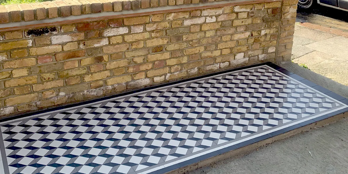 Side view of an Edwardian style mosaic tile pathway. The black, white and grey tiles make the optical effect of a cube pattern. Behind the path, an old yellow brick wall.