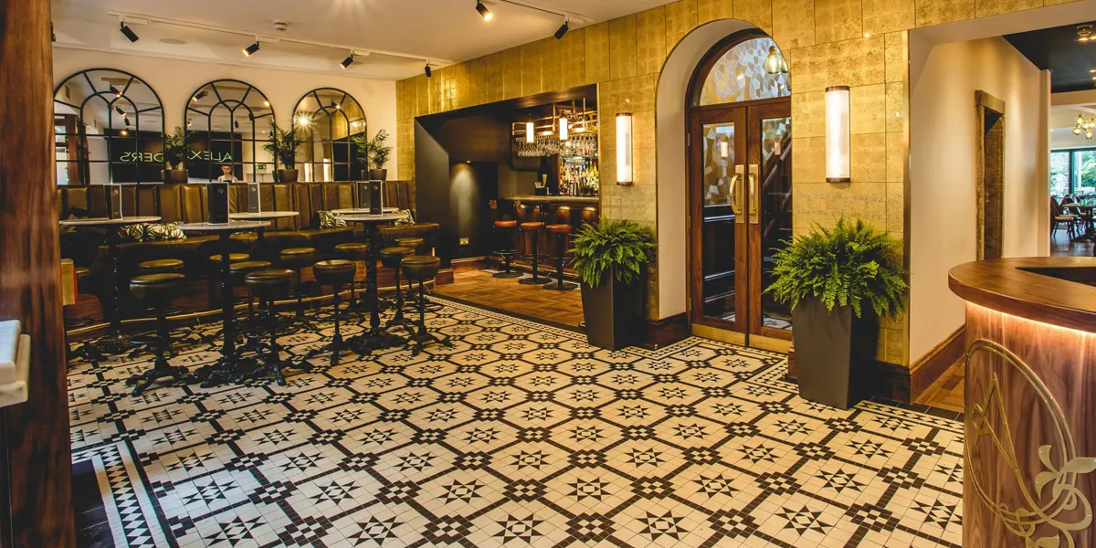 Black and white Georgian-style mosaic tiles seen on the lobby floor of a restaurant. There is a bar to the rear, and areas of seating with high stools. On each side of a wide wood and glass doorway, large square pots have green ferns growing.