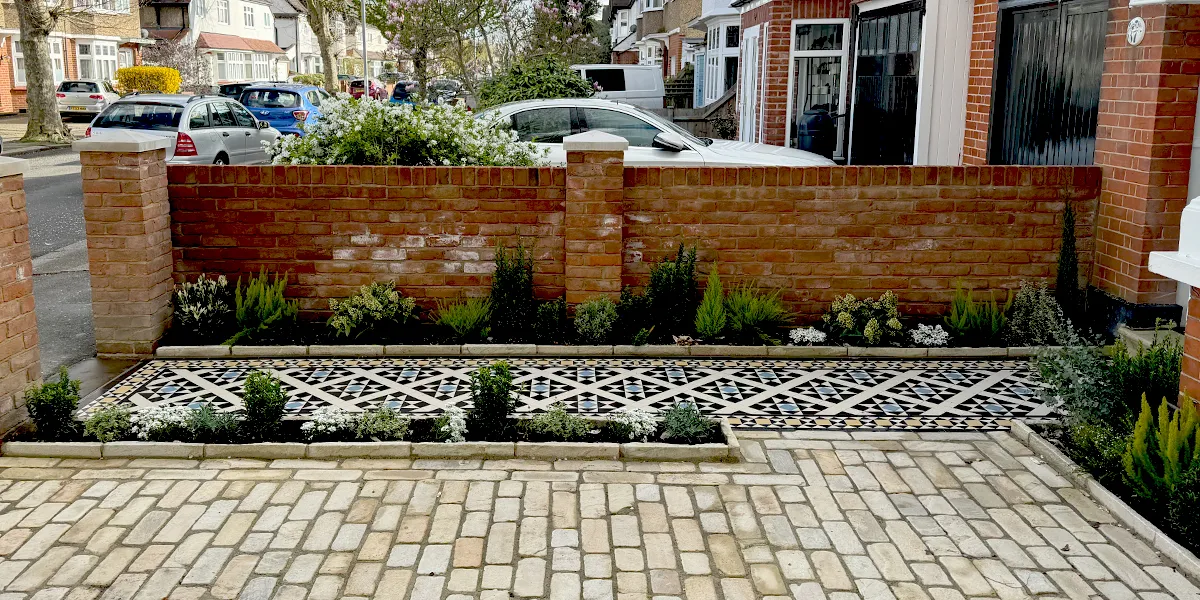 View from the side of a Victorian path mosaic in front of a red brick boundry wall with piers capped with stone. There are areas of planting to the edges of the path, and in front of the red brick house. The rest of the front garden is finished in stone. In the background a suburban street of semi-detached Edwardian houses is visible, there are several cars parked on the road and in driveways.