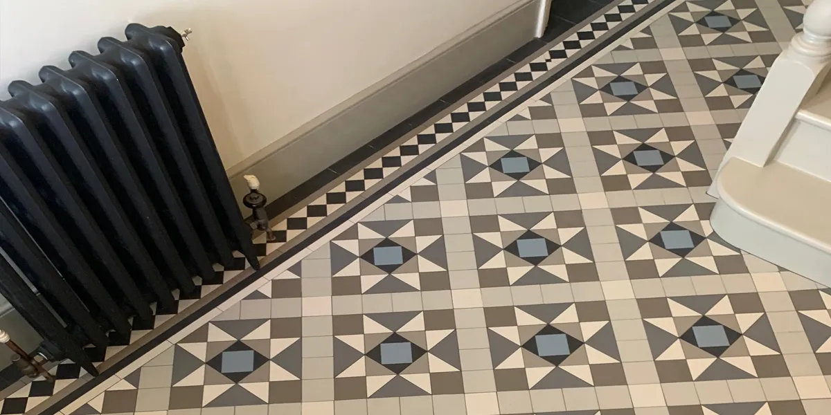 Looking down towards a Victorian tiled hallway floor, in black, white, grey and blue mosaic. To the left, a small black antique radiator, to the right, the base of a painted staircase.