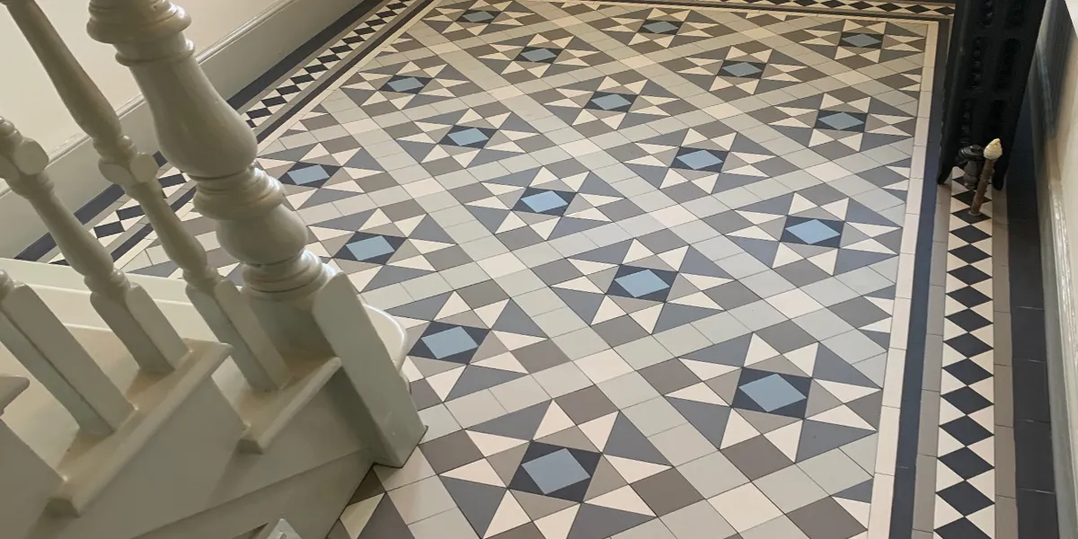 Victorian style mosaic hallway tiles in modern grey tones. The pattern is framed by a lines, and a black diamond border pattern. To the left, the base of a white painted staircase, with traditional spindles and newel post.