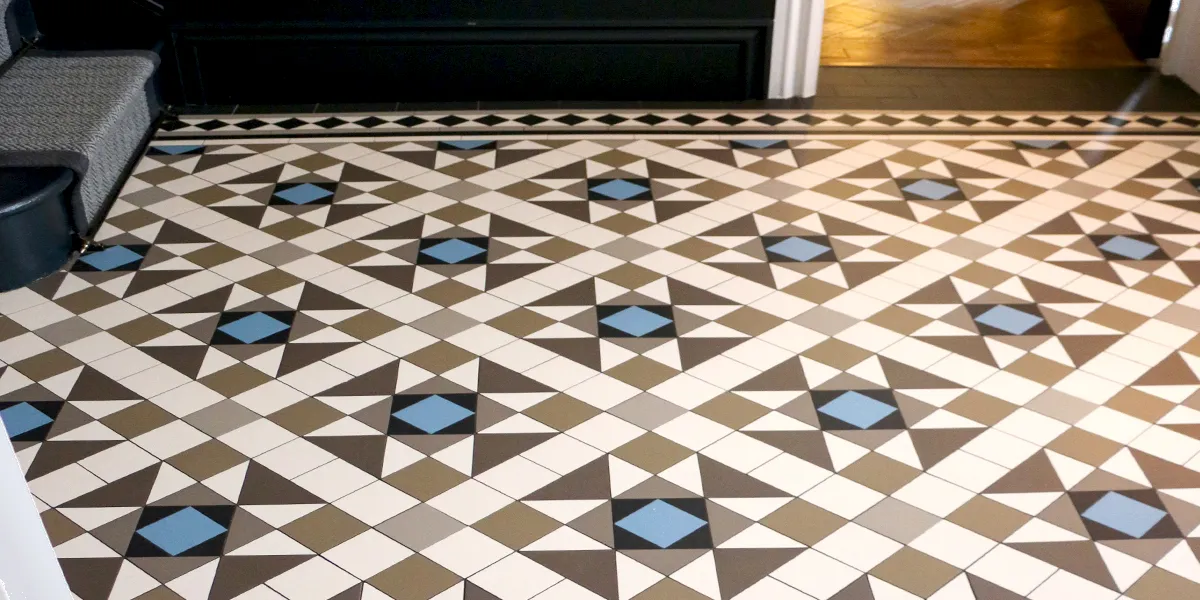 A closer view of a Victorian tile pattern in modern colours of greys and blue. The staircase and wall panels are painted black. A threshold to another room reveals wooden flooring.