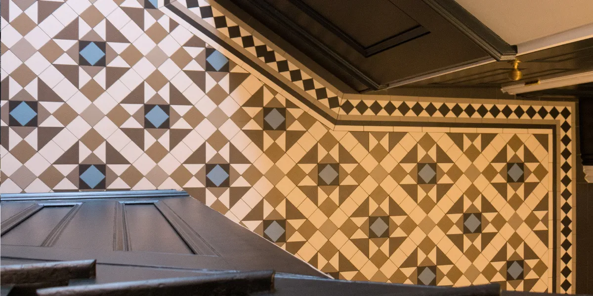 Looking down from halfway up the staircase at a modern coloured Victorian hallway mosaic. A traditional diamond border frames the design. The walls have wooden panels which are painted black, like the staircase.
