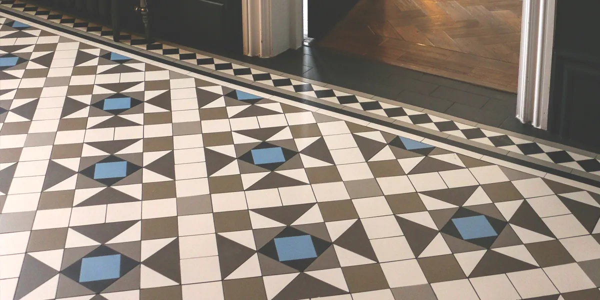 A close up view of a hallway mosaic in modern greys and blue tones. Natural light shines from the left. The wall panels are painted black, and the door architraves white. A threshold to another room reveals wooden flooring.