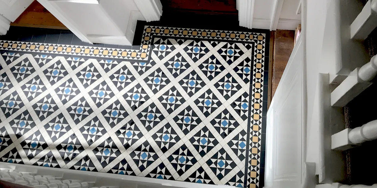 A view looking down at a Victorian mosaic hallway floor from the top of a staircase. The white gloss painted spindles are seen to the right and bottom. Three door thresholds lead to rooms with wooden flooring.