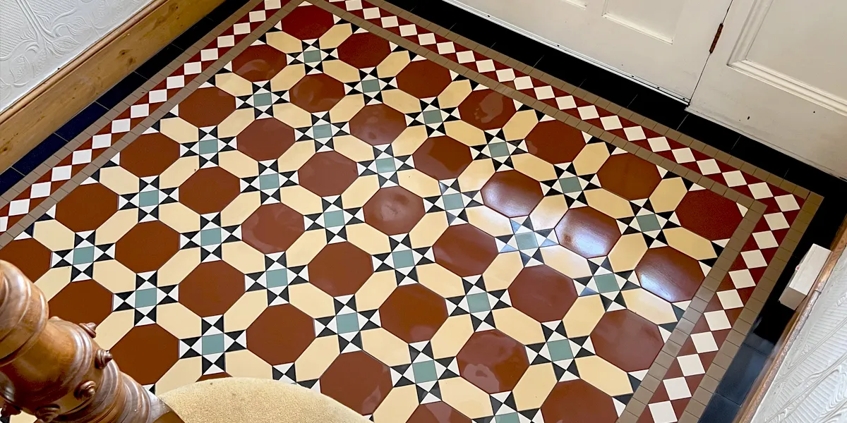 View looking down from a staircase at an Edwardian style mosaic hallway floor. The design is made with red octagons, long yellow hexagons, and a motif of small triangles and squares at the intersections of the pattern.