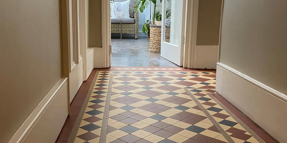 View of a coridoor area of a hallway, which has Edwardian-type tiles on the floor. There are tall, plain white skirting boards to the base of the walls, which are painted a light taupe shade. To the rear, a threshold leads to another room with large grey porcelain tiles on the floor and rattan furniture with cushions.