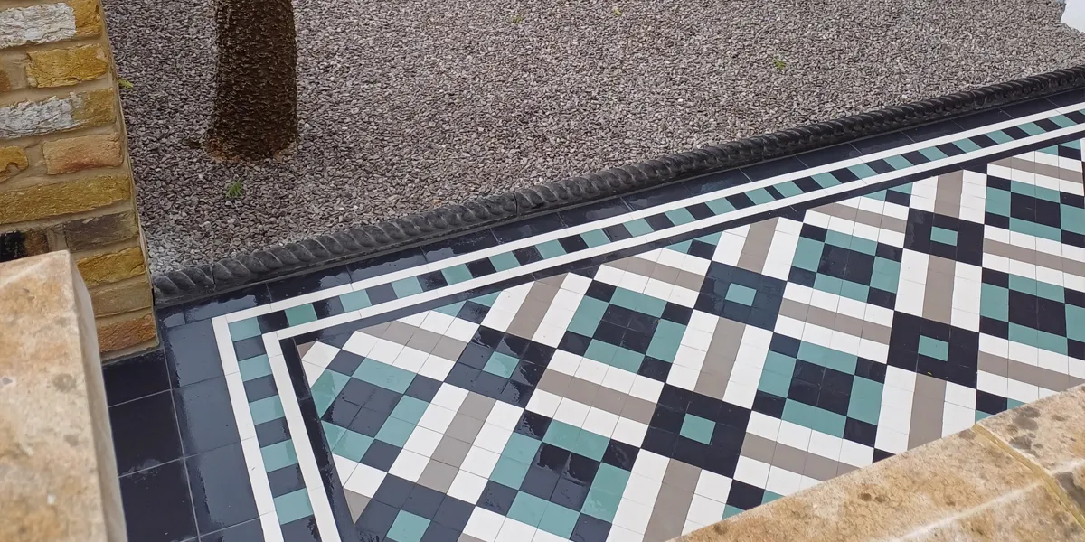 View from the side of a modern mosaic garden path, made of black, white, green and grey square tiles. The path is wet. The edge of the path has traditional ropetop, before the rest of the garden is gravel. To the left, yellow brick walls, with coping stones on top.