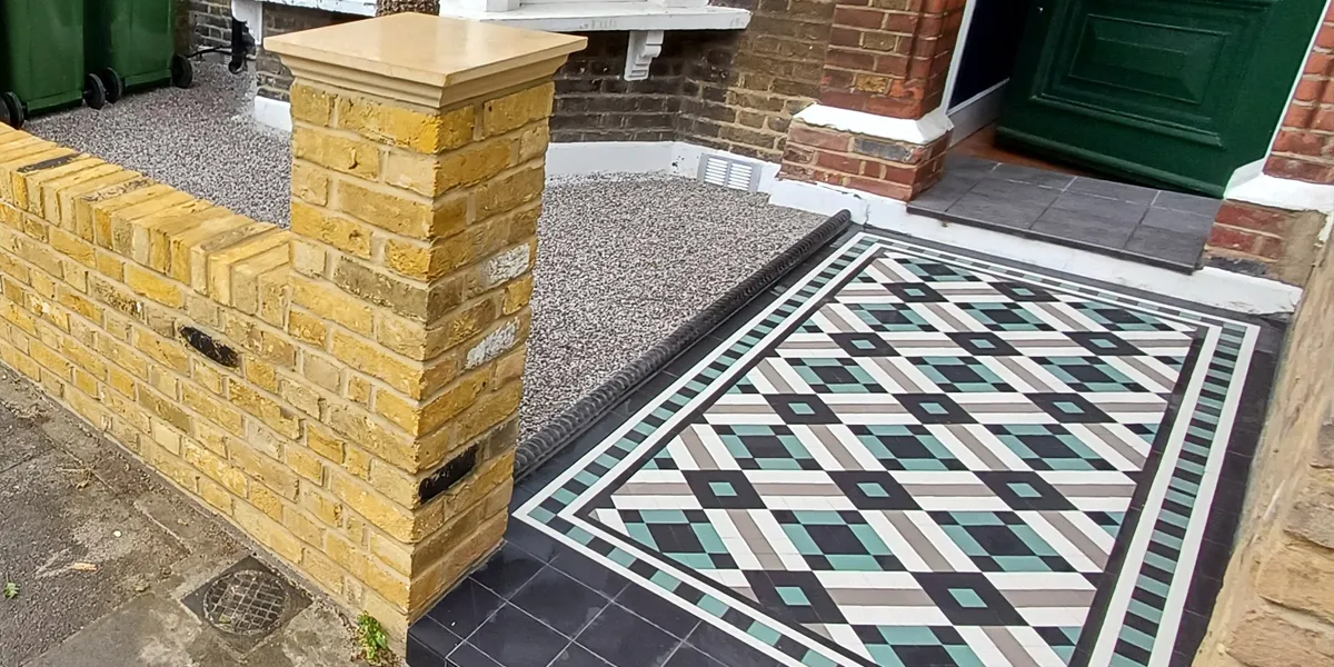 View across a front garden, with yellow brick front wall and gate piers topped with coping stone. A contemporary mosaic pathway leads to a small porch, with a deep green front door. The front garden is gravel.