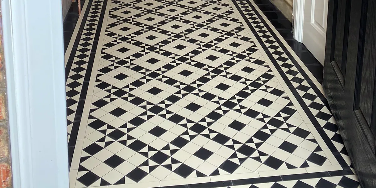 View through an open black door to a hallway tiled in detailed black and white mosaic. The design is framed with a border made of lines and a simple diamond motif.