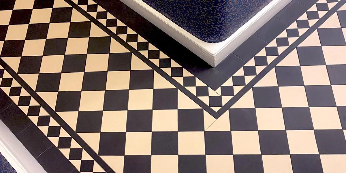The corner of a hallway with black and white checkerboard tiles to the floor. The wall has floral motif wallpaper, and the skirting boards are white.