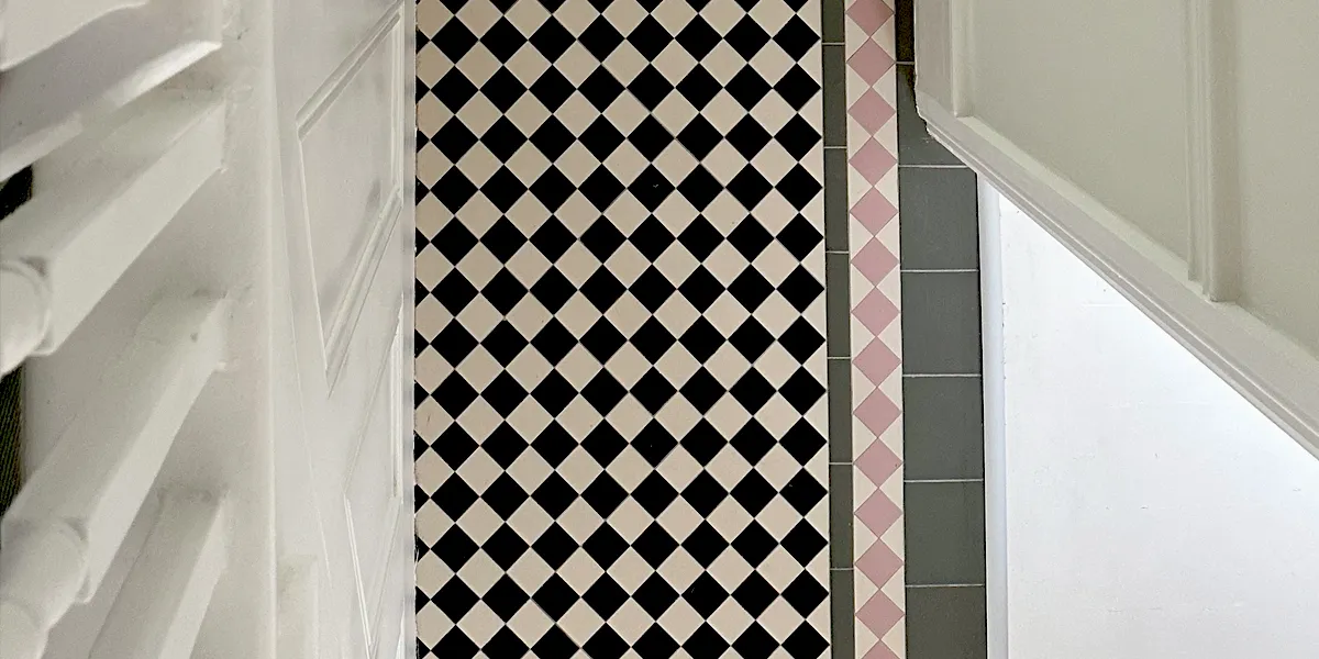 Birds-eye view of chequerboard tiled hall floor from side of staircase. Design features pink and green tiles in the border.