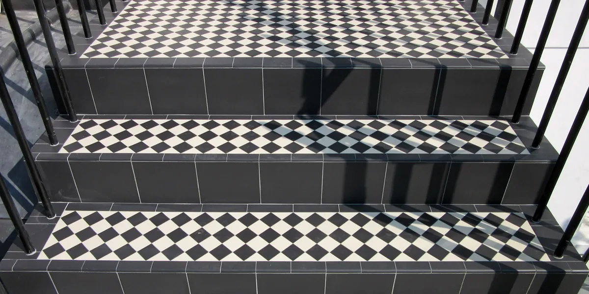 A set of steps with black and white chequerboard tiling to the treads, which are finished with round edge tiles. The step risers are larger black tiles. To the sides of the steps there are black metal railings.
