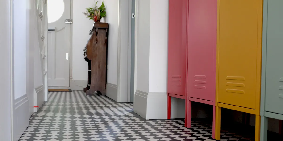 View from the back of a Victorian-style hallway with small black and white chequerboard floor tiles. To the back we see the front door with a circular window, and an antique wooden dresser. On the left, the side of the staircase, and to the right four metal lockers, painted in red, pink, yellow and blue.