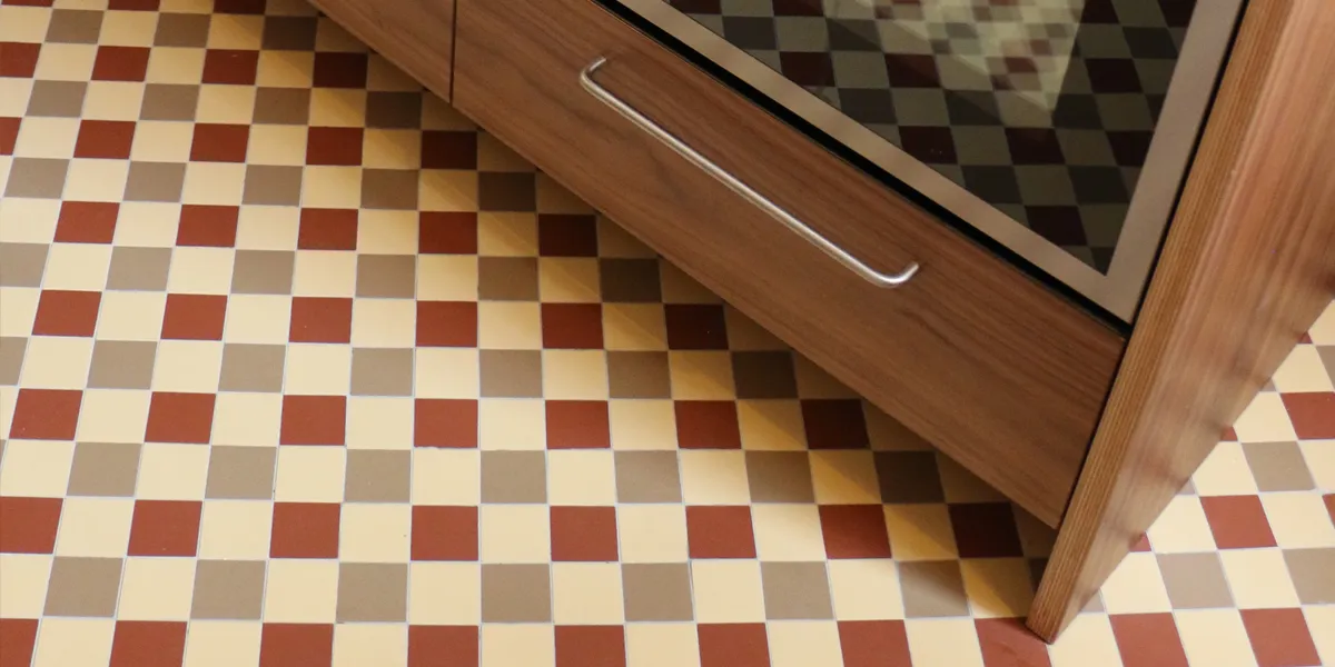 Tri-coloured chequerboard floor tiles fitted in a kitchen. The kitchen cabinets are a dark wood, and the over door reflects the mosaic floor.