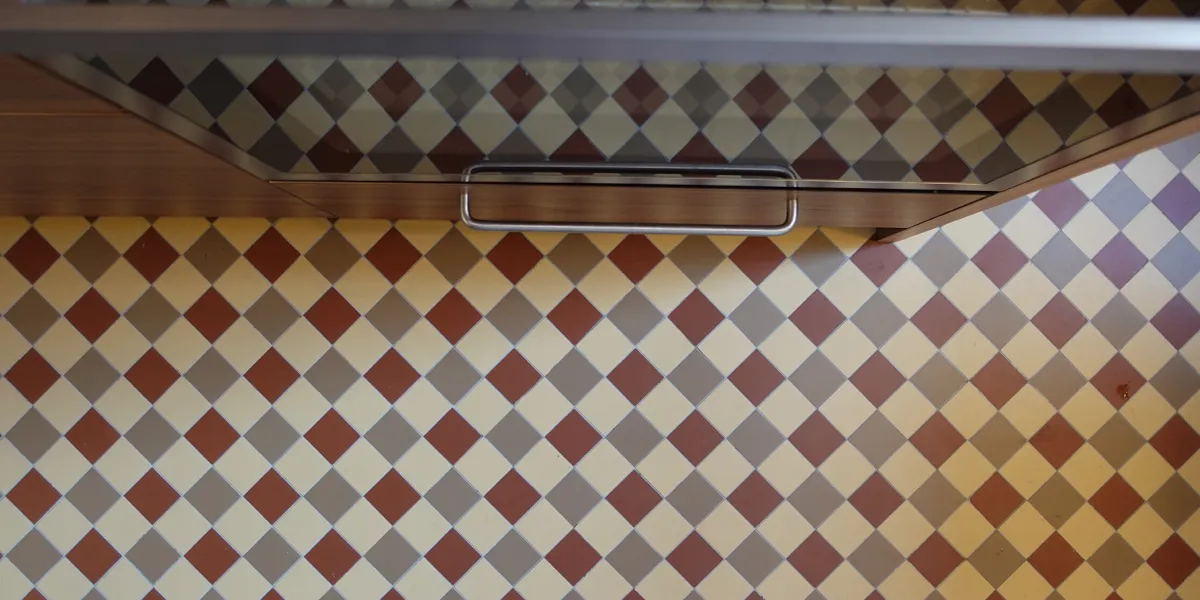 View looking down at a mosaic floor made of red, buff and oatmeal coloured tiles. To the top a glass oven door reflects the floor tiles. The kitchen cabinets are a dark wood tone.