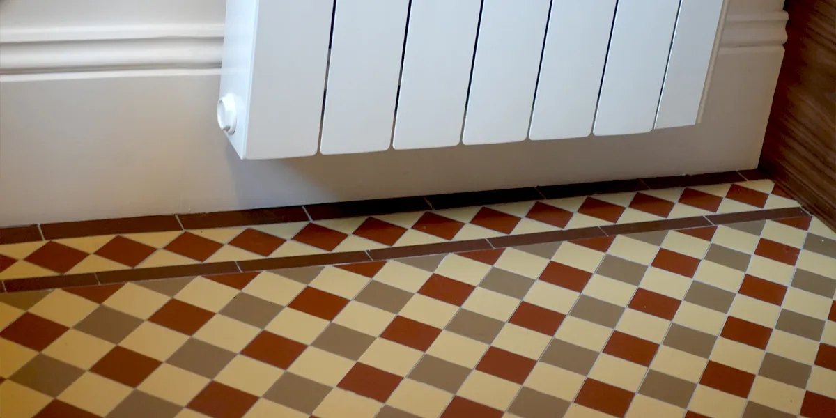 Close up view of a three colour chequerboard mosaic on a kitchen floor. The border is a diamond pattern against a large white skirting board. A white radiator is partially seen to the top of the picture.