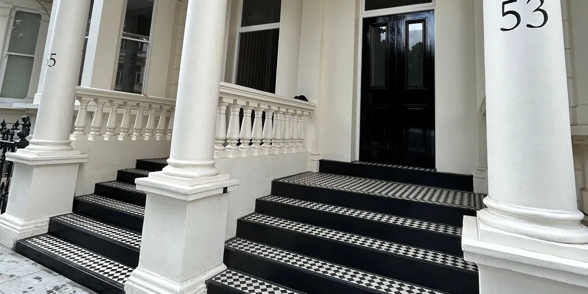 Two sets of steps tiled in black and white mosaic leading to a Regency period townhouse with large gloss black front doors. There are three large columns at the foot of the steps, and stone balustrades to the sides - both rendered and painted white. Number 53 is seen at the top of the right-hand column.