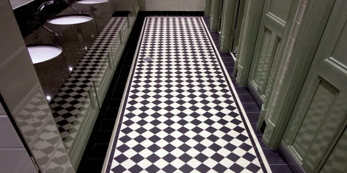 Black and white Victorian-style mosaic tiles fitted to the floor of a public house toilet. To the left several wash-basins set within a black-speckled top. To the right, doors to private toilets are painted a sage green colour. The floor tiles can be seen reflecting on the vertical surfaces alongside.