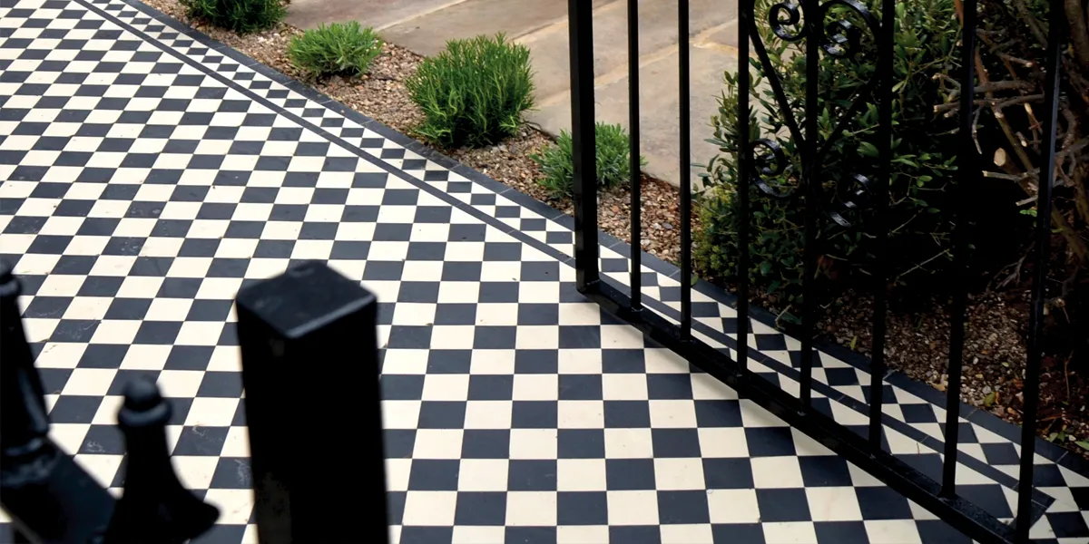 A view of Victorian black and white pathway tiles leading towards the left. The design uses larger tiles centrally and smaller tiles in the border. The garden has wrought iron railings and gate. To the right of the chequerboard path a small area of planting with gravel base, then stone slabs to the rest of the garden.