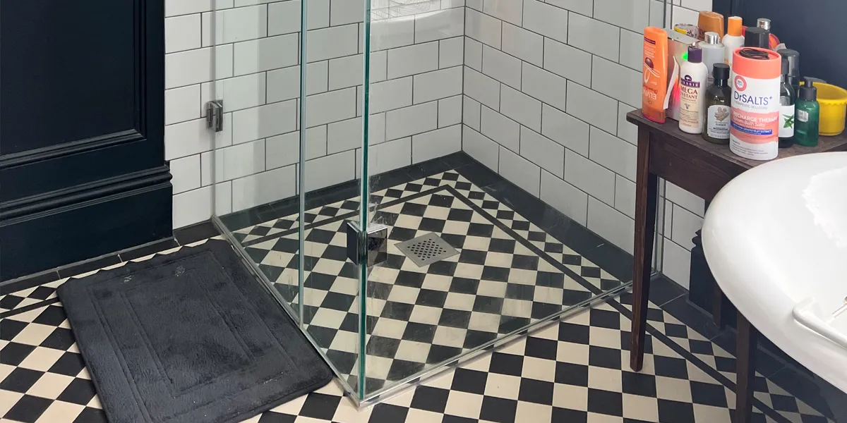 Black and white Victorian chequerboard tiles on a bathroom floor. A glass panel shower area has a metal drain in the center, a dark grey mat sits in from of the shower. To the right a small wooden table with shampoos and conditioners.