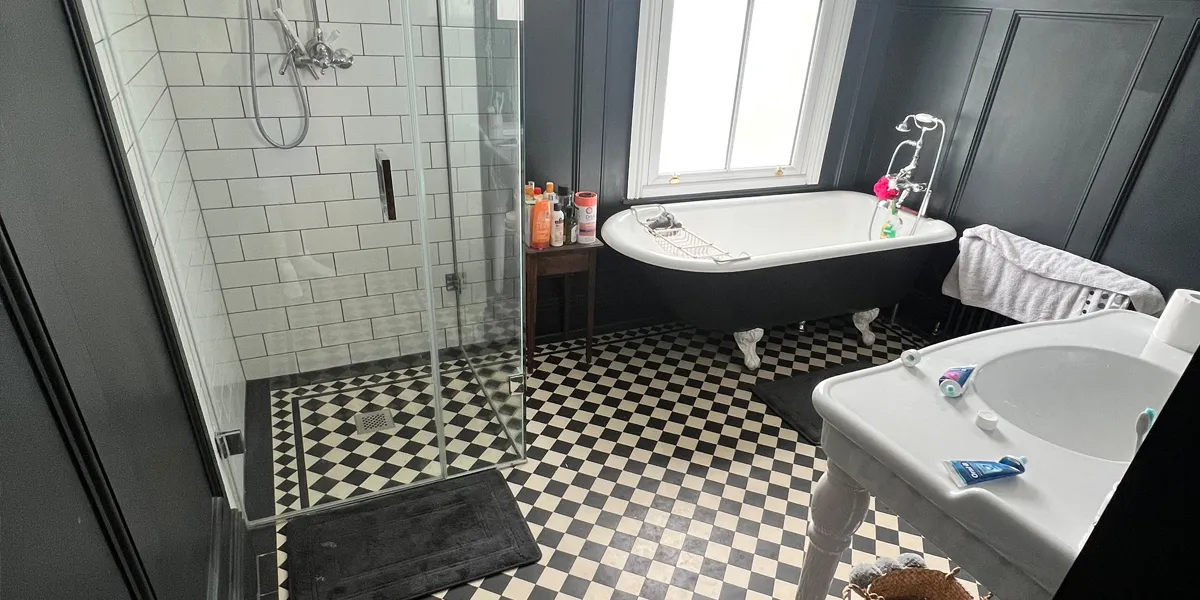 A spacious Victorian type bathroom with natural light flooding through a large window. The walls have wooden panelling painted black. The floor has traditional black and white chequered tiling. There is glass panelled shower, a free-standing bath with shower fitting, and large white sink with turned legs complete the look.