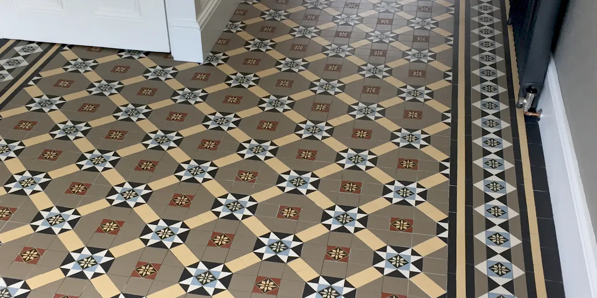 Traditional and highly decorative, multi-colour Victorian encaustic tiles on a hall floor. The design is composed of a trellis pattern with star motifs. The back of the staircase is on the left, and to the right a black cast-iron radiator.