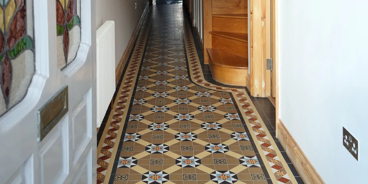 A multi-coloured Victorian encaustic hallway mosaic runs in to the distance. To the right, a wooden staircase rises. To the left, a white painted front door with stained-glass inserts is open.