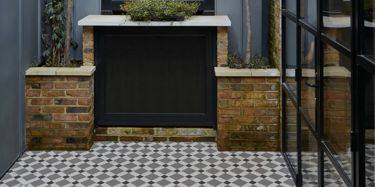 Black, white and grey geometric mosaic tiles across a patio garden. Red brick planting area to the rear, and glazed doors leading inside to the right.