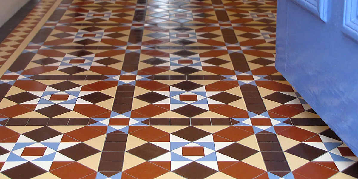 Traditional Victorian hallway tiles ob a hall floor. The design is multi-coloured and uses a combination of squares and triangles in a tessellating pattern. To the right, the bottom of a bright blue front door.