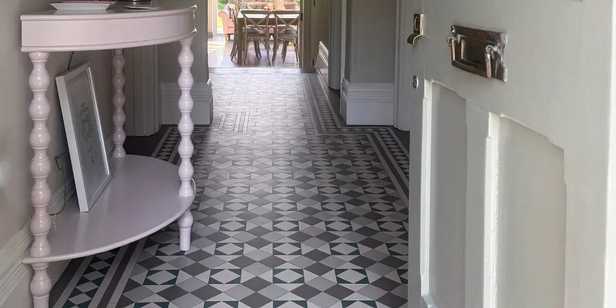 View through an open, white painted front door, of a hallway in a Victorian style property. The floor has a mosaic tile pattern in a modern colour palette of greys and green. To the left, a dusty pink painted three legged table. To the rear of the hall there is a threshold to a dining room and sofa sits in front of patio doors.