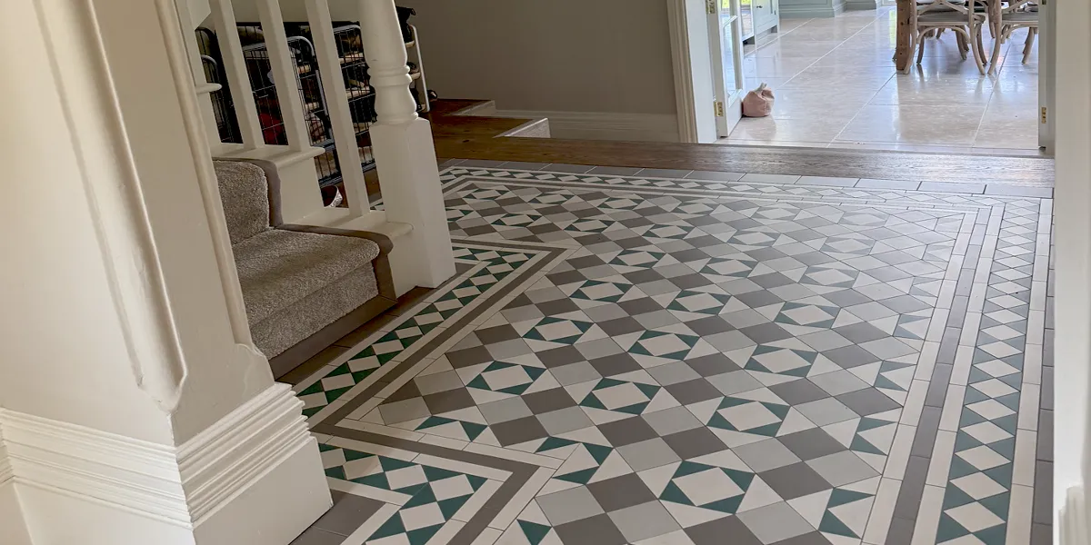 A traditional type of mosaic tile pattern in modern colours decorates a hallway floor. To the left a staircase with beige stair-runner. The rear of the hallway stes down and a wide doorway leads to a kitchen.