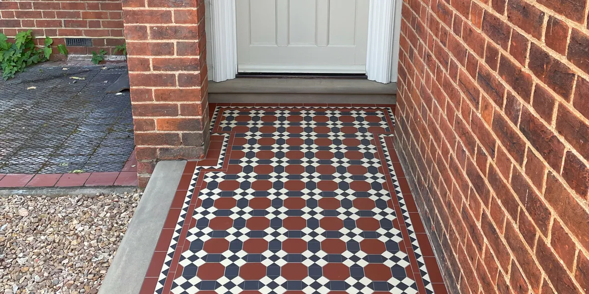 Red, black and white Edwardian style mosaic path tiled design featuring octagons between a tresllis of square flower motifs, leads to a beige front door. To the left and right a burnt red brick wall. To the left of the path a stone edging leads to an area of gravel.