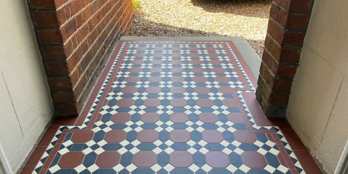 View from a porch area looking out along an area of Edwardian-style mosaic tiles. The tiled area is finished on two sides by stone edging, which leads to a gravel front drive lit by sunlight. The house has red bricks, and the inside of the porch walls are painted white.