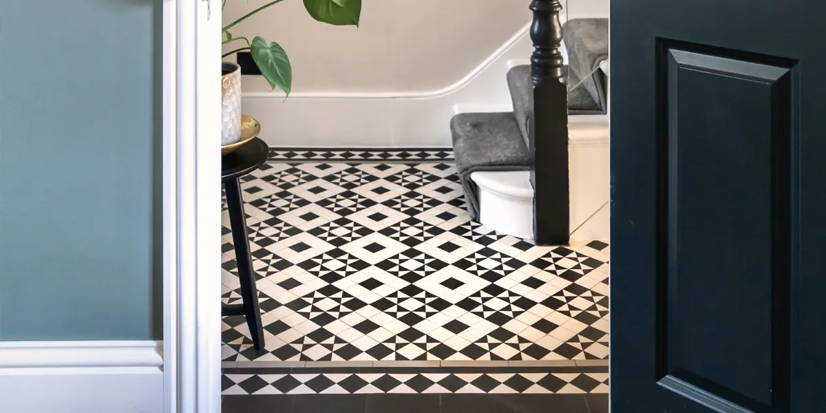 View through an open black door of a hallway with black and white Victorian-style mosaic tiles. The pattern contrasts a square against a star motif. The staircase and skirting boards are gloss white, and there is a grey stair-runner. To the left a small stool has a potted plant on top.