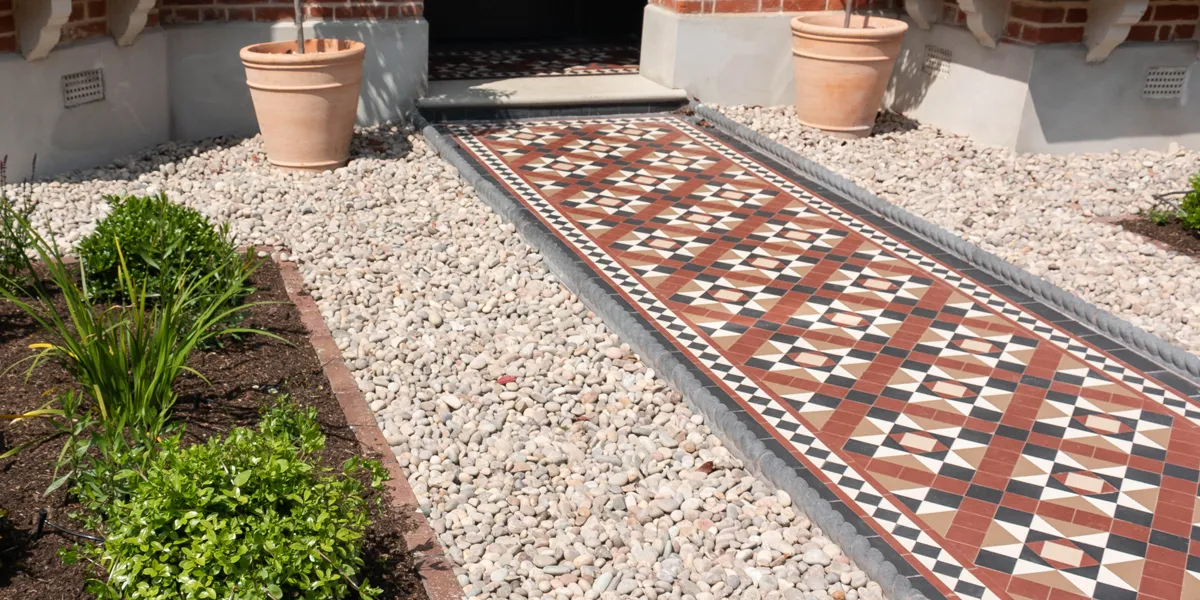 An Edwardian style mosaic pathway in red and black tones leads to a stone porch step. To the right and left, Victorian-style rope top edging holds back areas of pebbles. To the left and right there are new areas of planting with small shrubs. To the left and right of the porch step are terracotta pots.