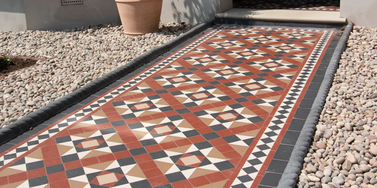 A traditional mosaic pathway design using an Edwardian style pattern in reds and browns. The path is finished with ropetop edging to the sides. The rest of the front garden has a deep layer of different sized pebbles. The bottom of a single terracotta plant pot can be seen in front of the base of the house.