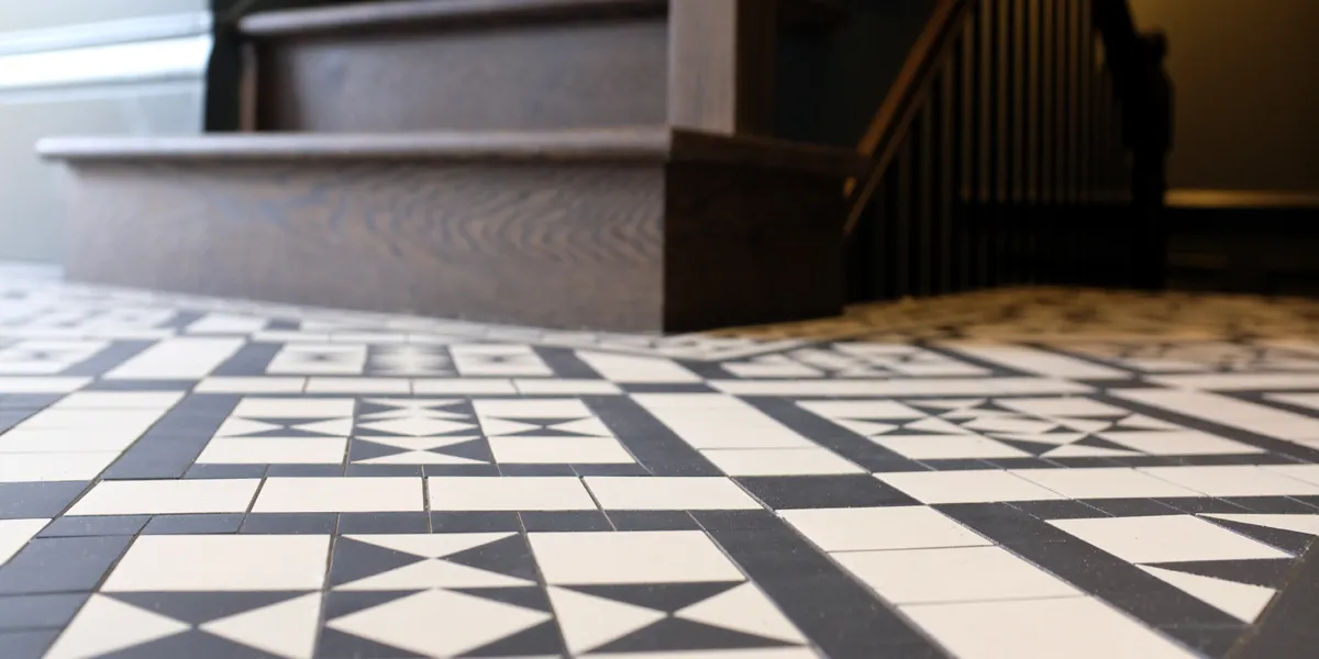 Close up view of black and white mosaic tiles in a hall. A deep brown stained wooded staircase leads upwards and also down to the basement.