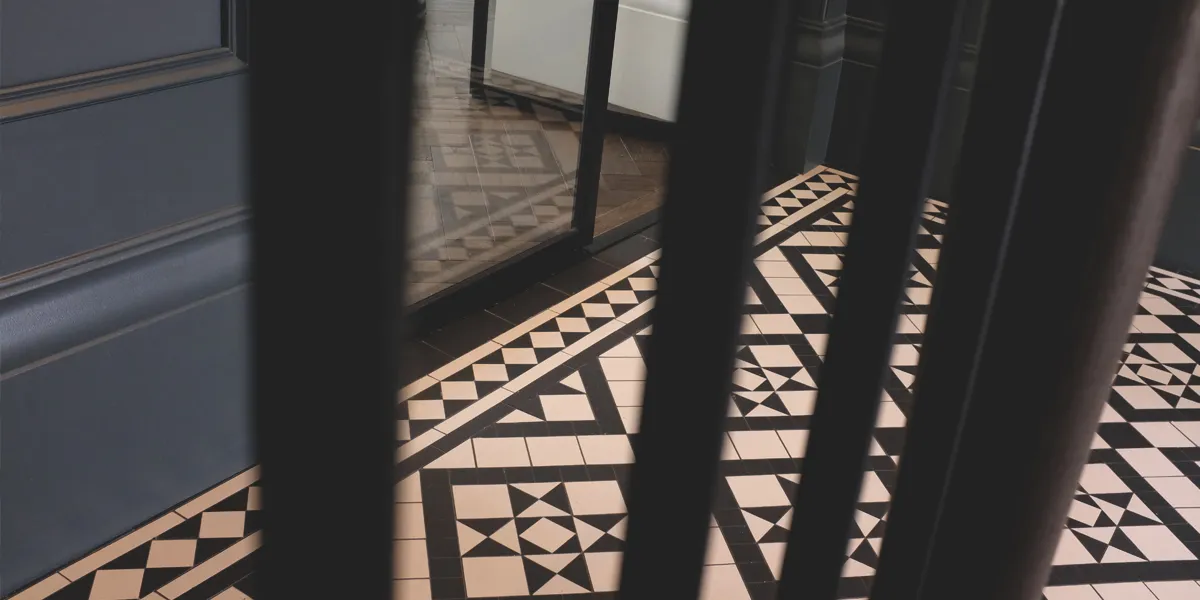 A Georgian mosaic floor tile design in a hallway viewed through staircase spindles. Glass doors leading to a parquet floor reflect the tile design.