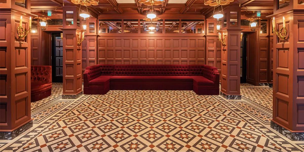 A hotel bar with Victorian-style mosaic floor which follows the features of the space - highlighting thresholds and different areas of the bar. There is moulded timber panelling, and a coffered ceiling with layered lighting. To the rear a large wrap around, red velvet, banquette.